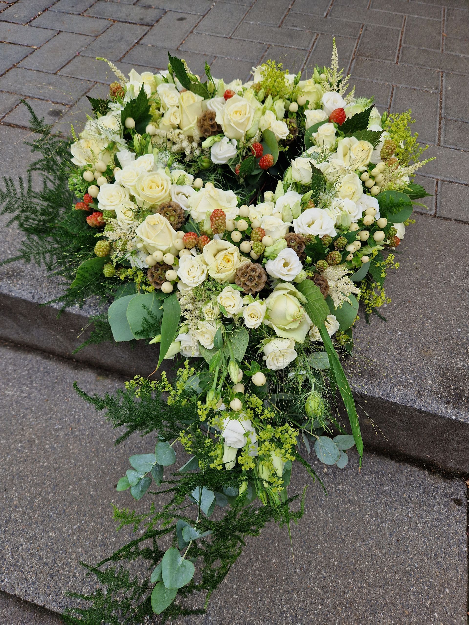 Auf einem Gehweg liegt ein herzförmiger Kranz aus weißen Blumen und Grünpflanzen.