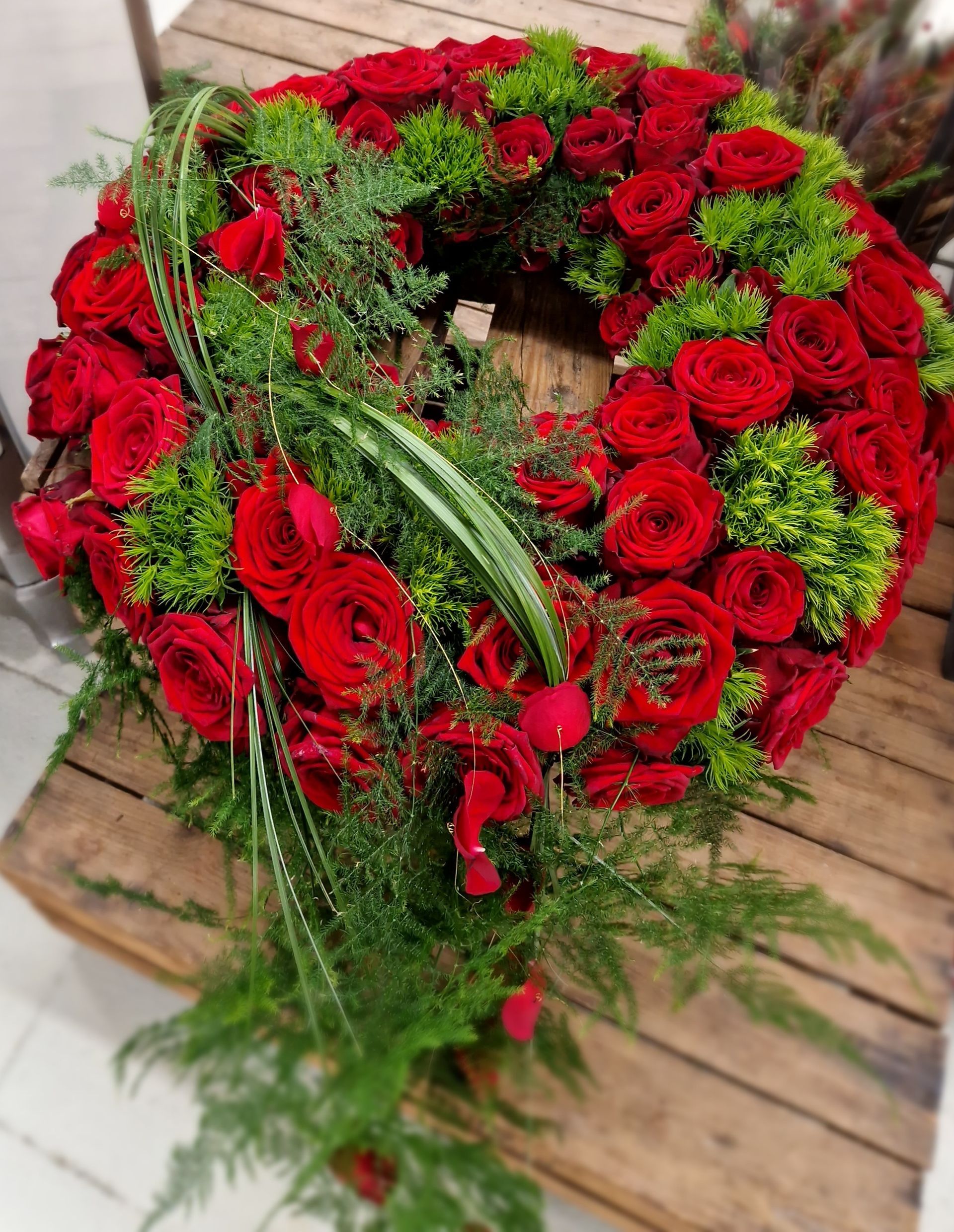 Ein Kranz aus roten Rosen und Grünpflanzen steht auf einem Holztisch.