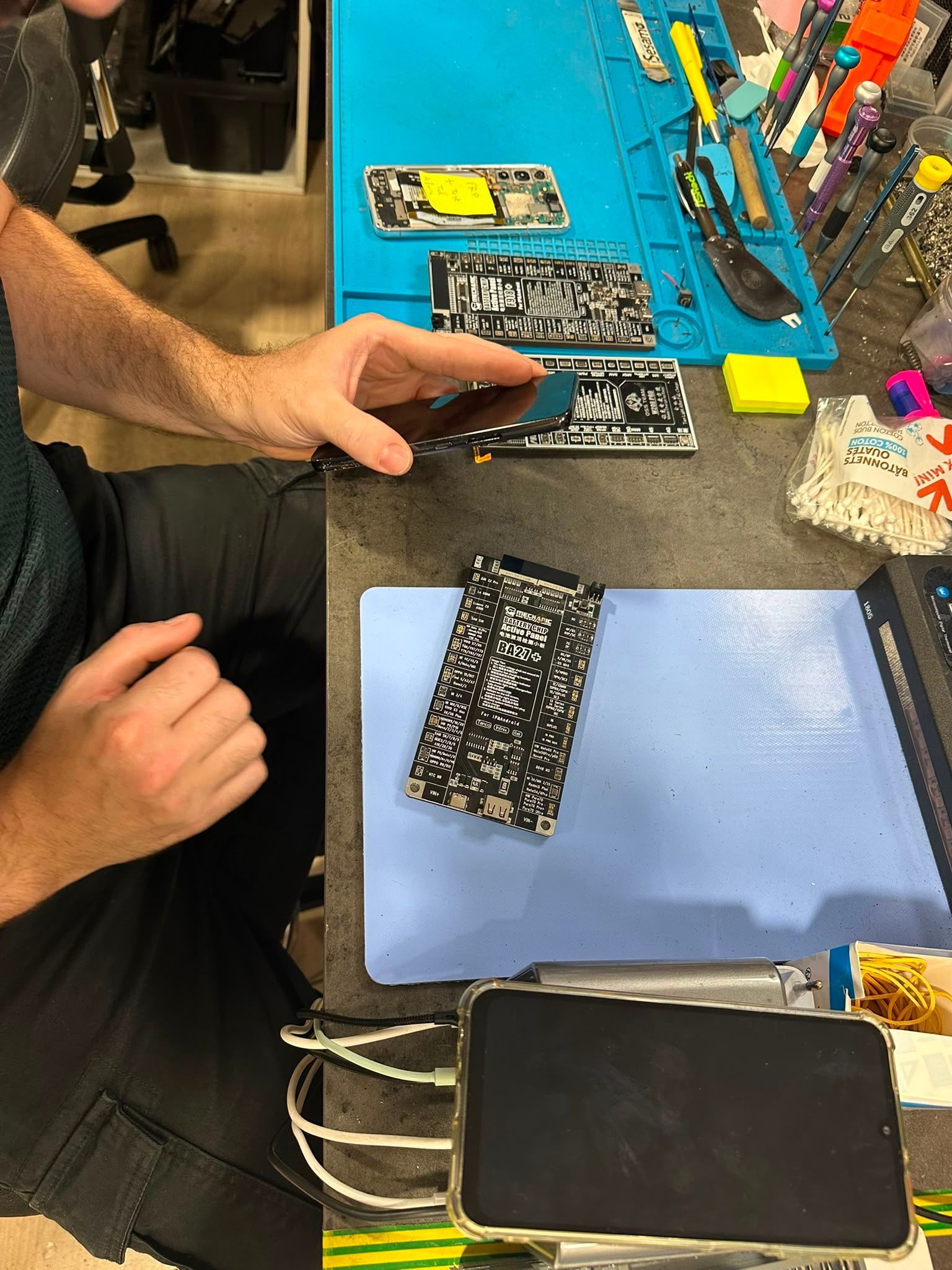 Une personne manipule un écran de téléphone brisé sur un établi de réparation, avec des outils et des pièces à proximité.