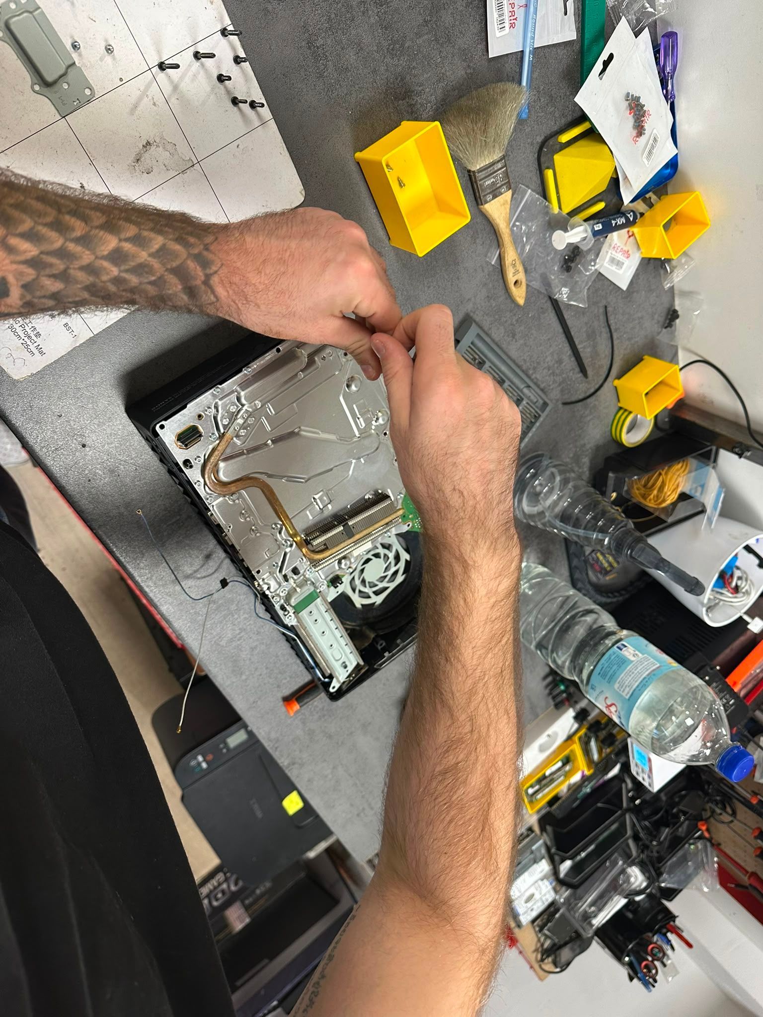 Des mains réparent les composants internes d'un ordinateur portable ouvert sur un établi, avec des outils et des composants électroniques à proximité.