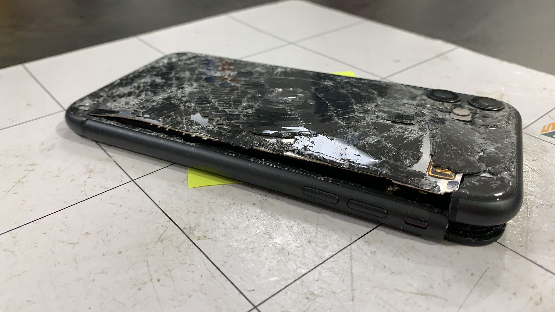 Un smartphone noir fissuré, posé face contre terre sur un sol carrelé, avec un petit mot jaune en dessous.