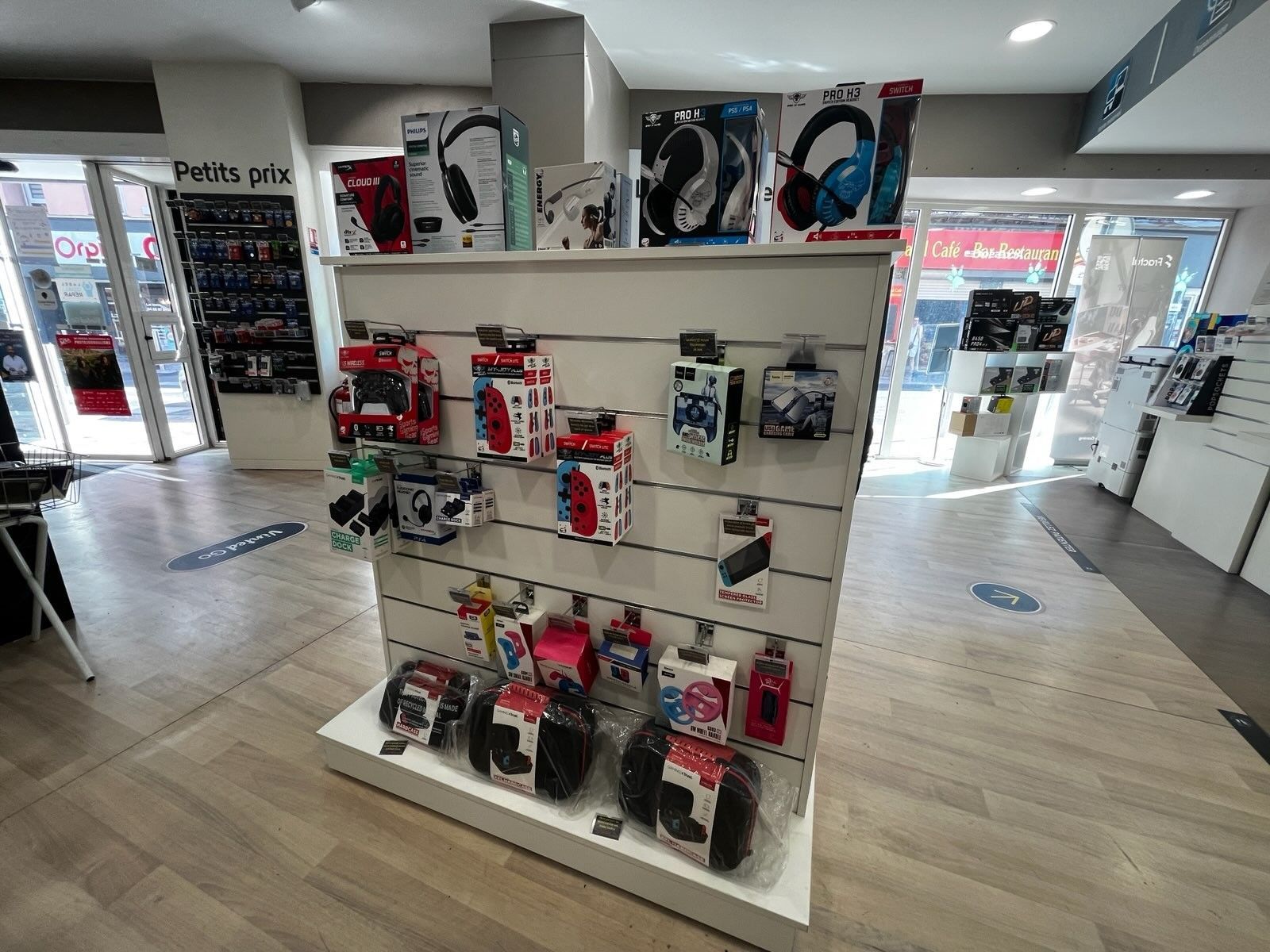 Présentoir de casques audio et d'accessoires emballés sur des étagères blanches dans une allée lumineuse d'un magasin d'électronique.