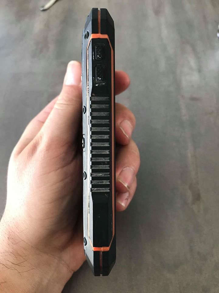 Vue latérale d'un smartphone noir robuste avec des accents orange, tenu dans une main