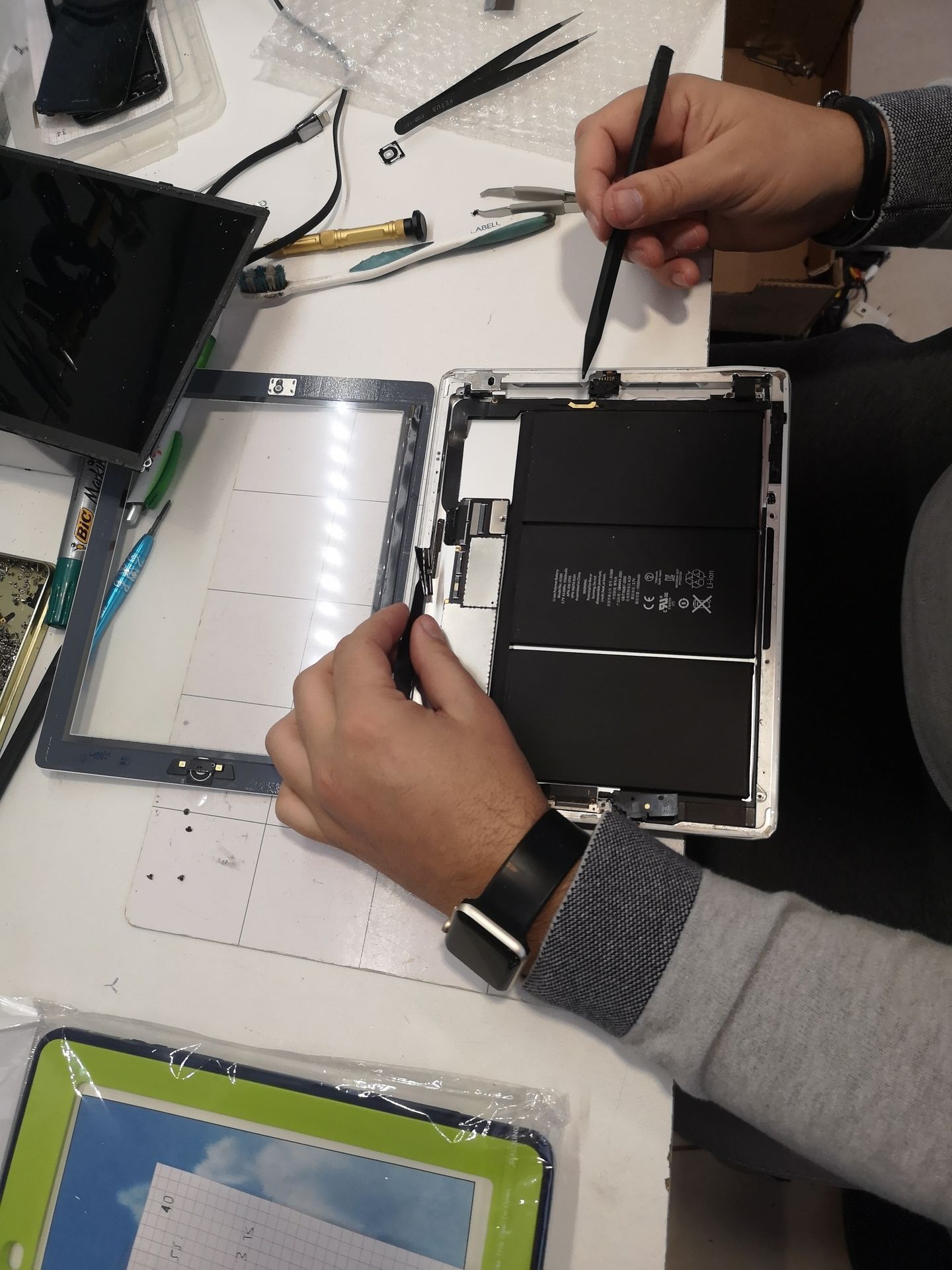 Des mains réparent une tablette ou une liseuse noire sur un établi, des outils et des pièces détachées étant éparpillés à proximité.