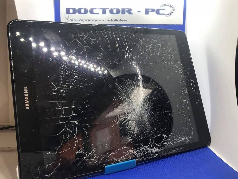 Tablette noire fissurée à écran brisé, posée sur un support bleu sous une enseigne Doctor-PC.
