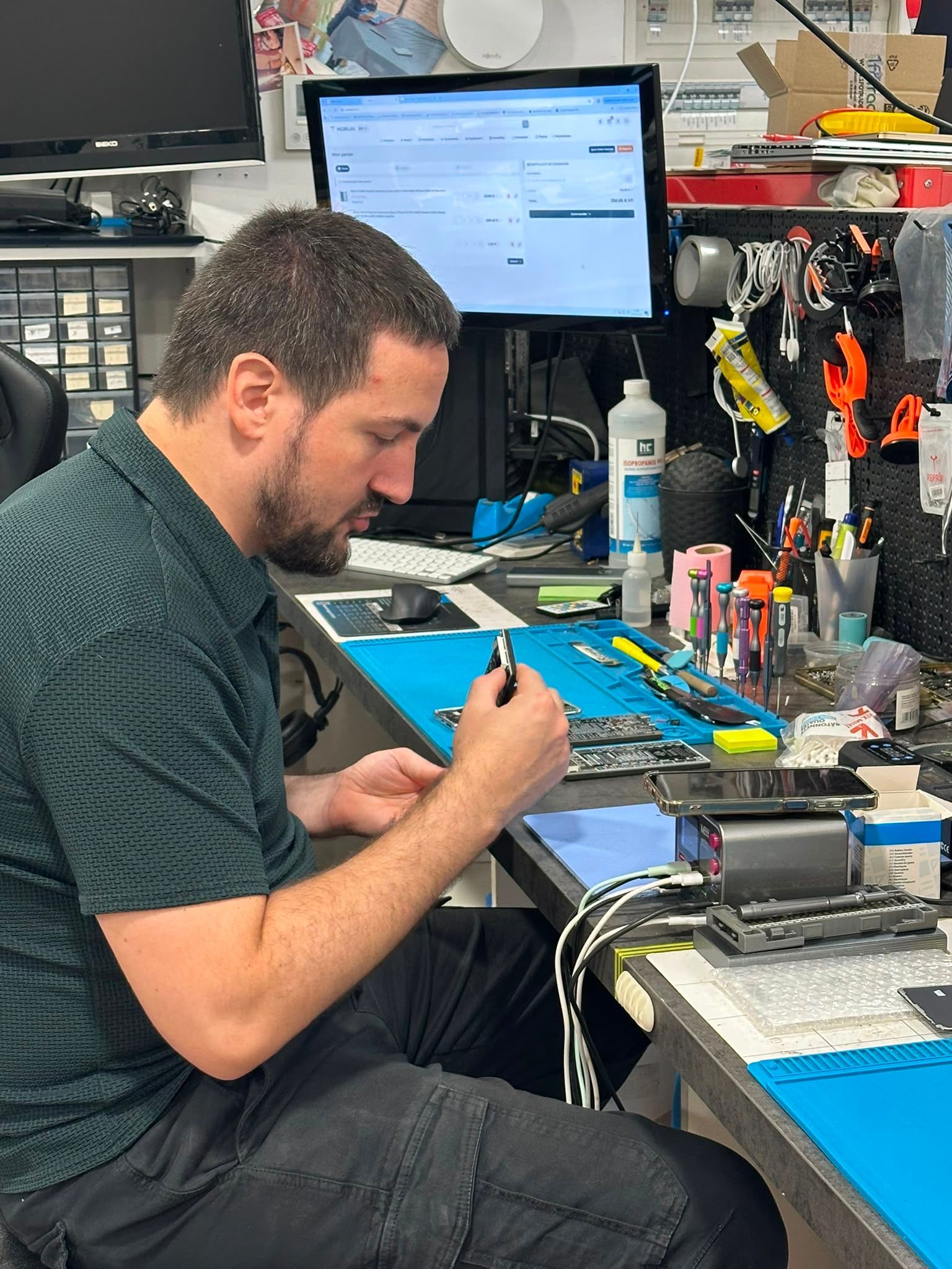 Un technicien travaille sur un établi de réparation électronique encombré, inspectant un petit outil ou appareil.