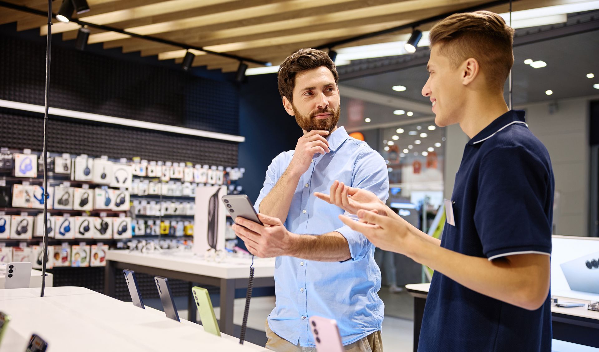 Deux hommes discutent dans un magasin d'électronique, l'un tenant une tablette et l'autre faisant des gestes.