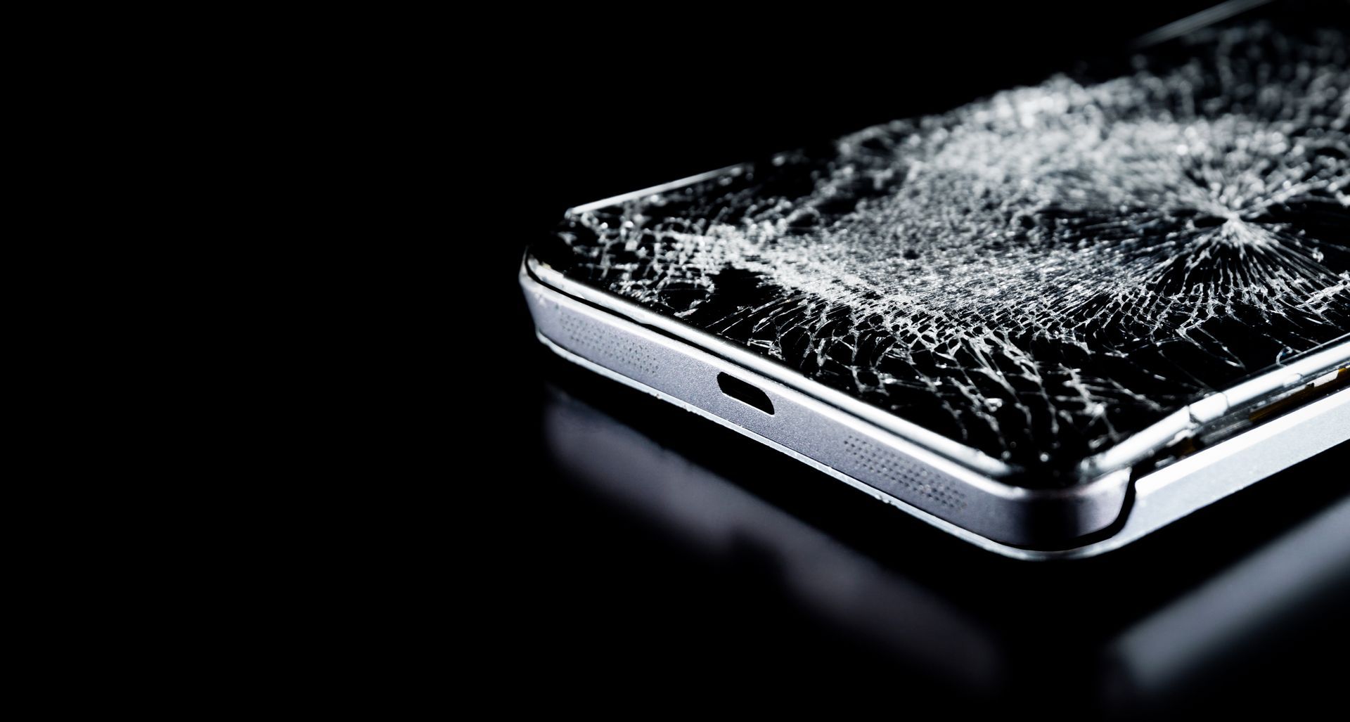 Écran de smartphone fissuré sur fond noir, montrant des éclats de verre et l'extrémité du port de chargement.