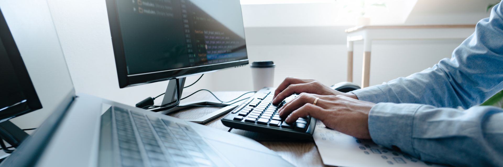 Des mains qui tapent sur un clavier numérique à côté d'un ordinateur portable et d'un écran dans un espace de travail de bureau lumineux.