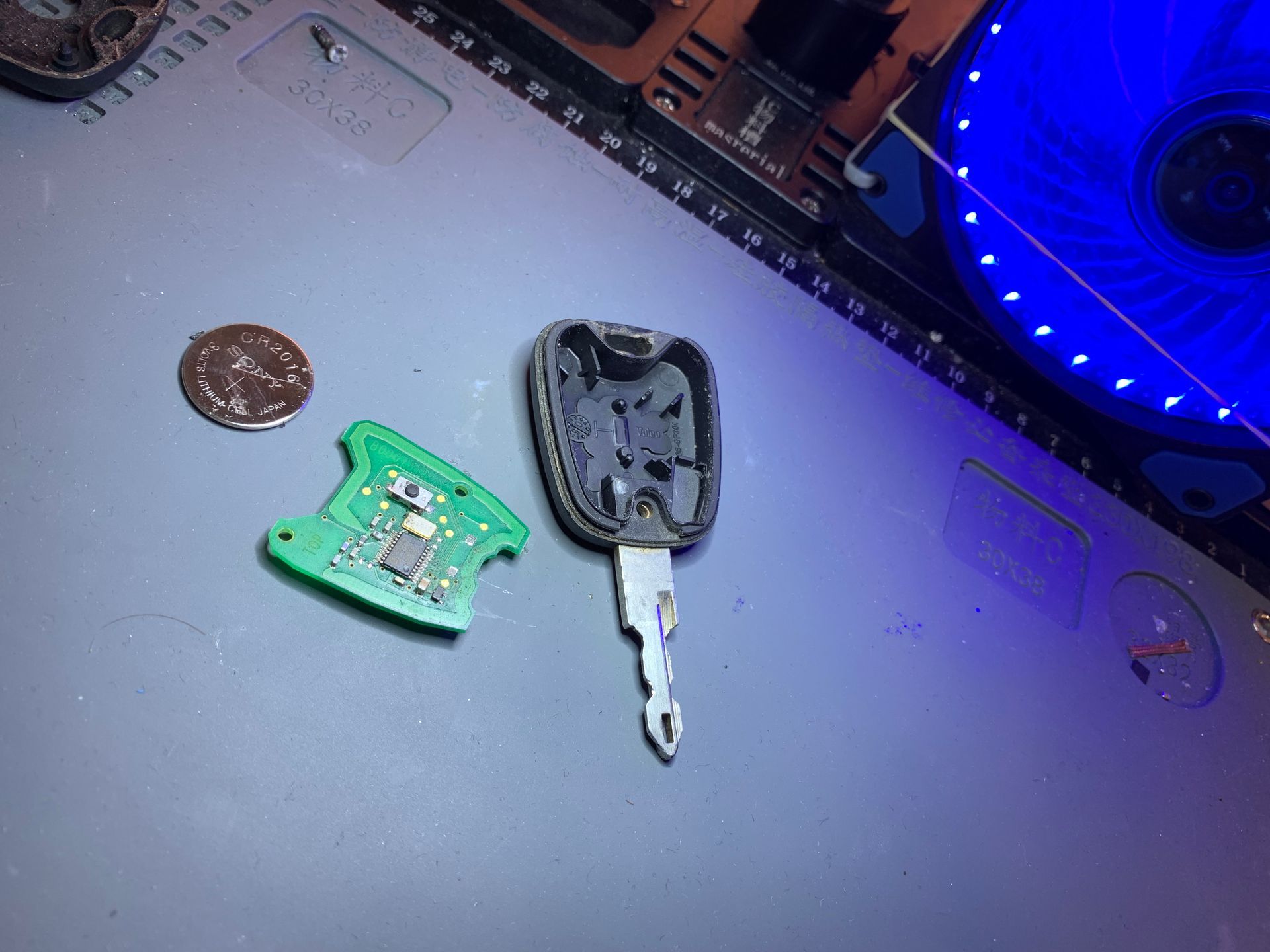 Un porte-clés noir cassé et des morceaux de circuit imprimé vert sur une surface grise près d'un ventilateur à lumière bleue.