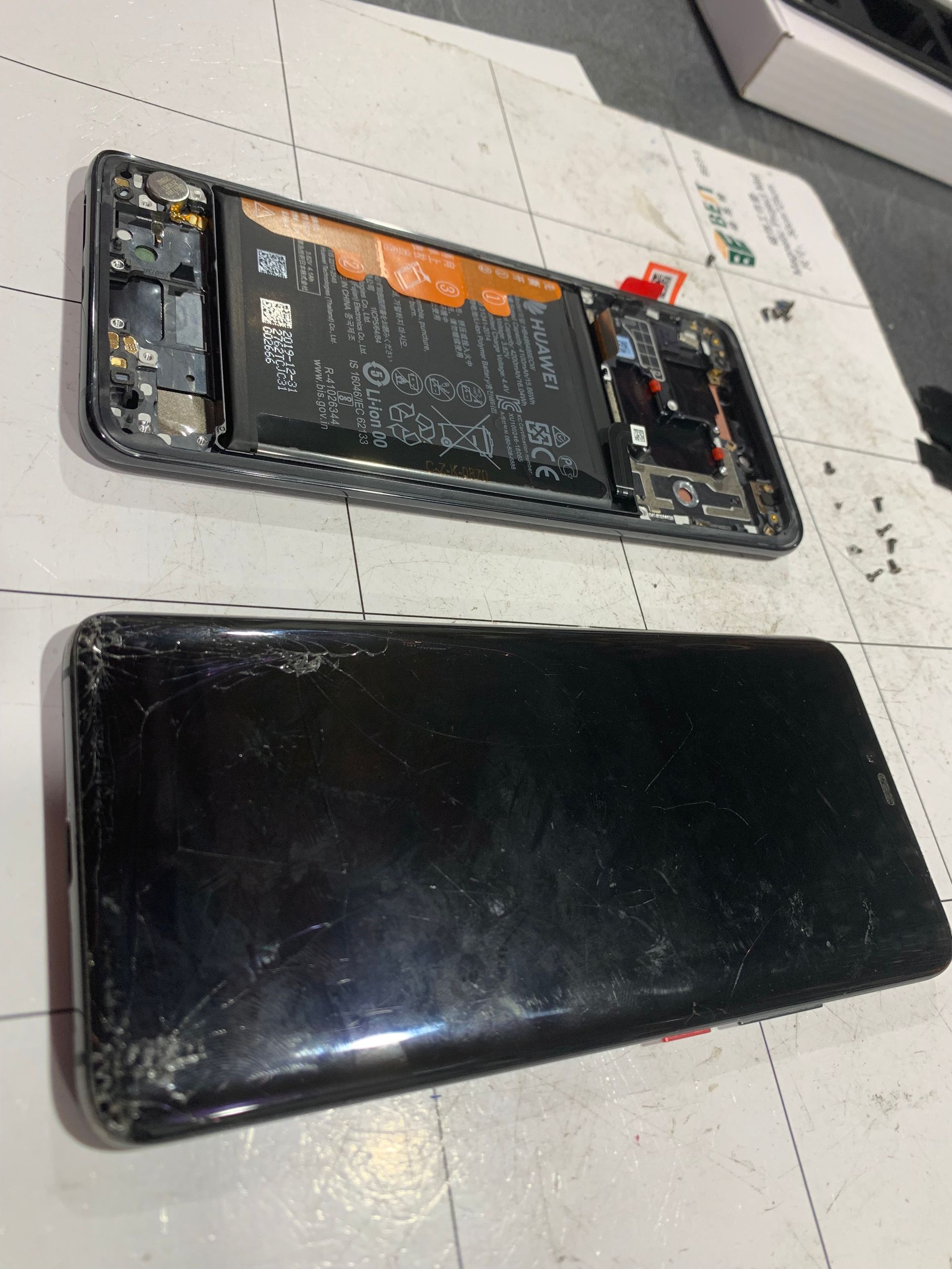 Smartphone démonté, batterie apparente, au-dessus d'une coque arrière noire, sur un sol carrelé.