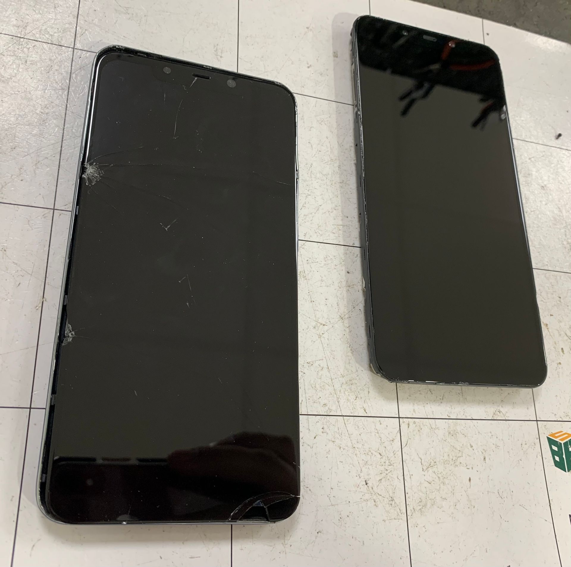 Deux smartphones noirs, l'un plus grand, l'autre plus petit, sont posés face visible sur un sol carrelé.