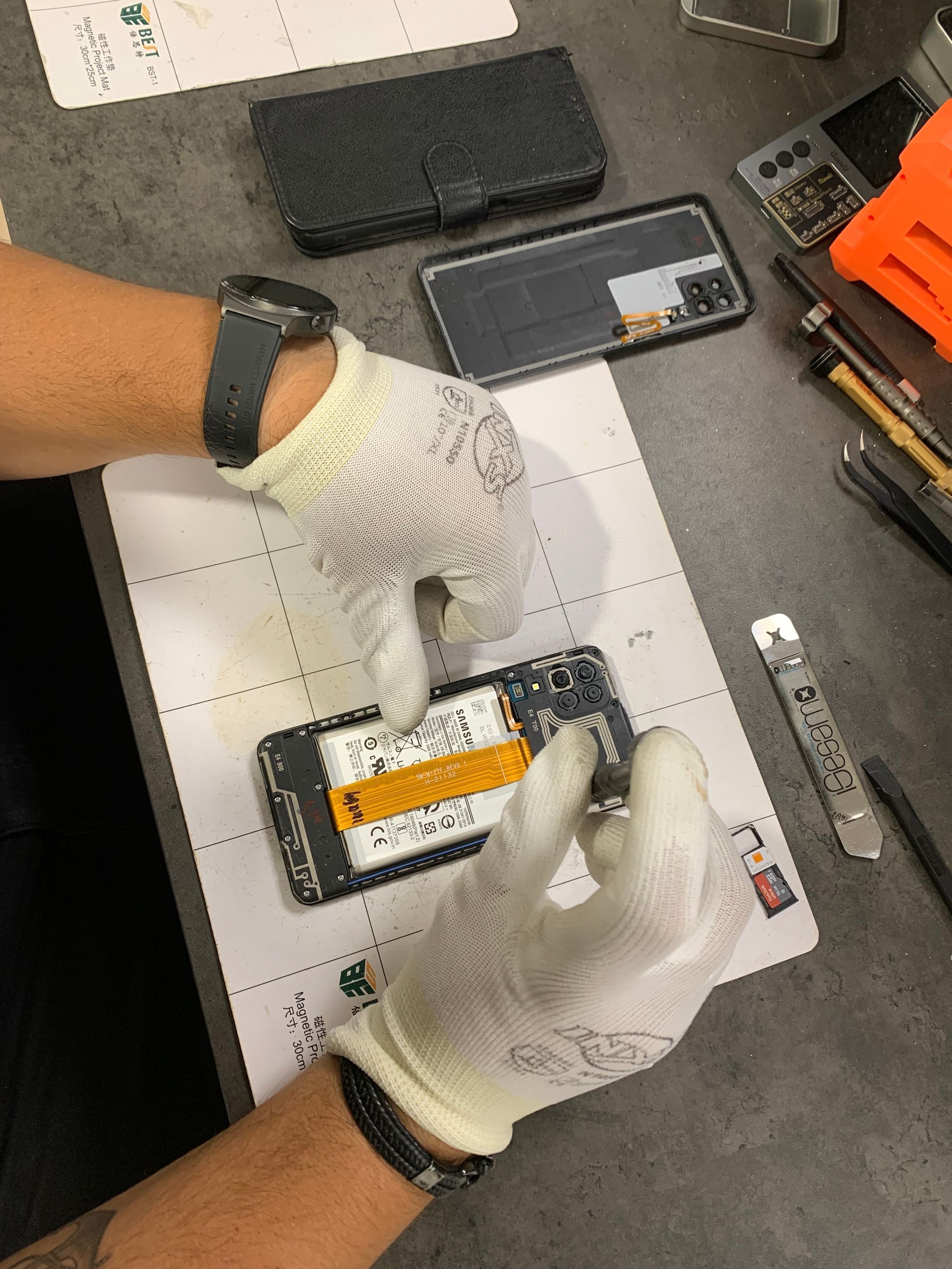 Des mains gantées inspectent une batterie de smartphone sur un établi, à proximité d'outils et de pièces détachées.