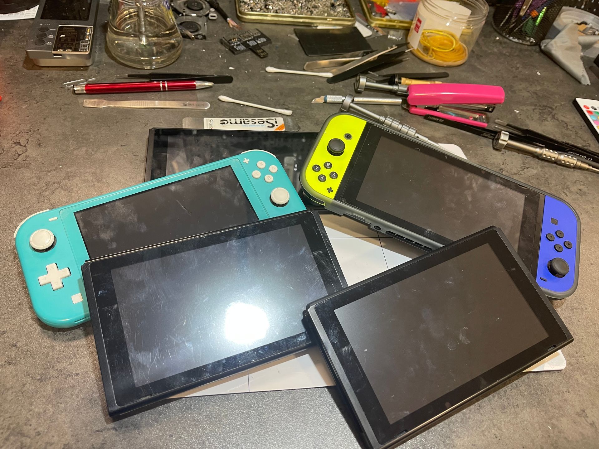 Trois consoles de jeux et deux tablettes sur un établi encombré, avec des Joy-Cons colorés et des outils à proximité