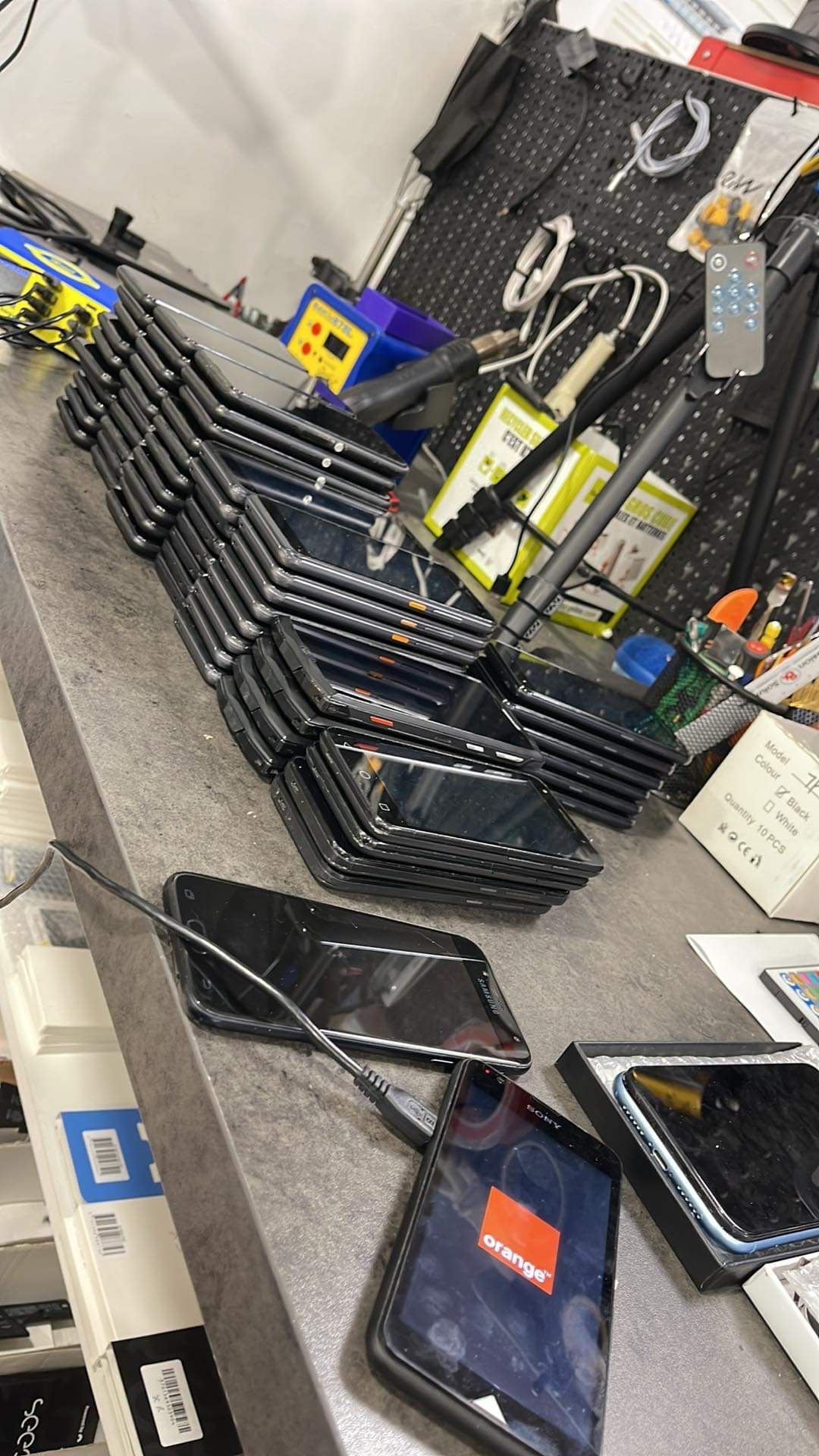Des caisses à outils et des sacs à outils noirs empilés sur le sol d'un atelier, à côté d'une table où sont disposés outils et pièces détachées.
