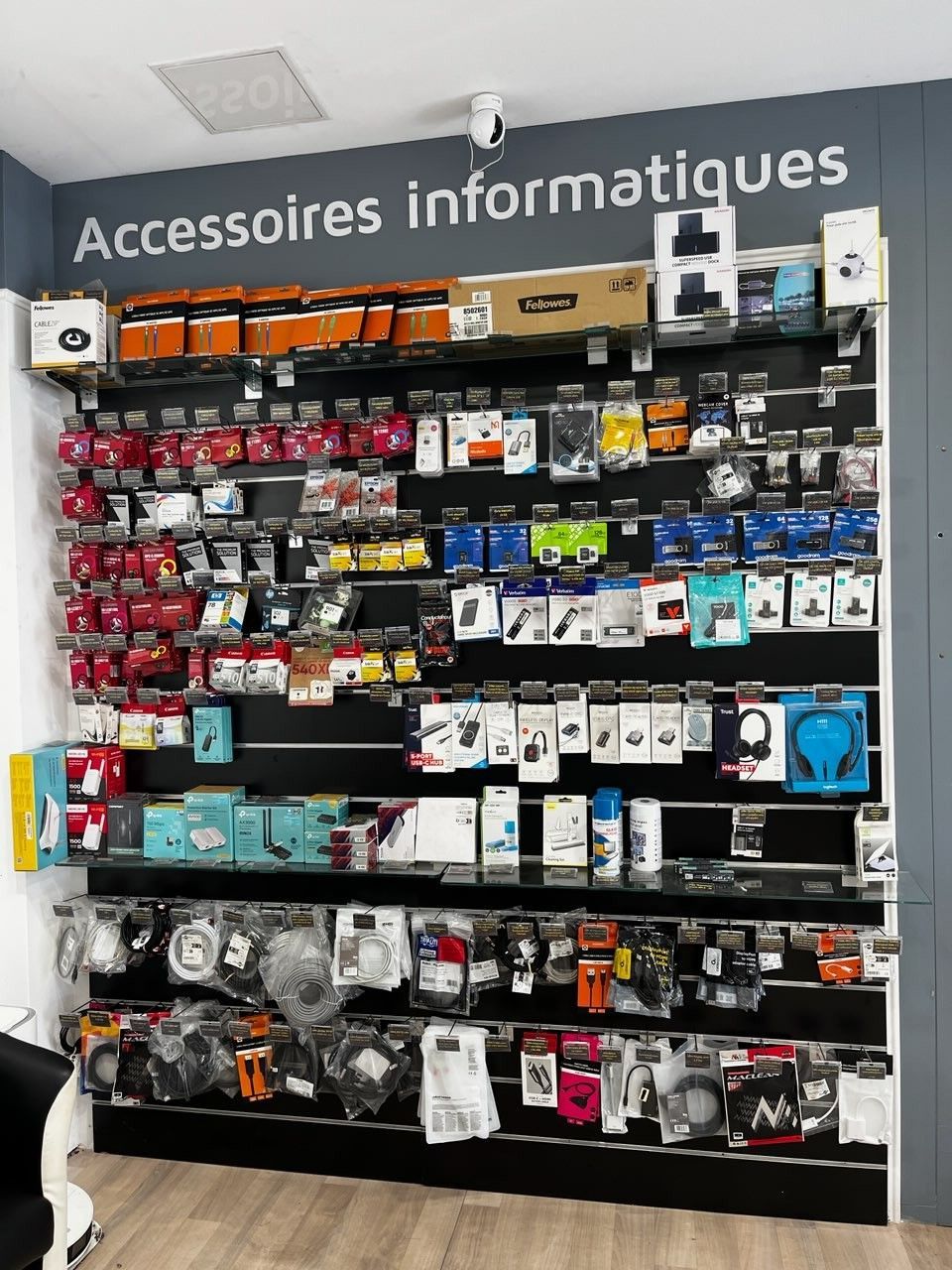Mur d'accessoires informatiques dans un magasin, avec des câbles, des adaptateurs et des gadgets emballés assortis sous l'enseigne « Accessoires informatiques ».
