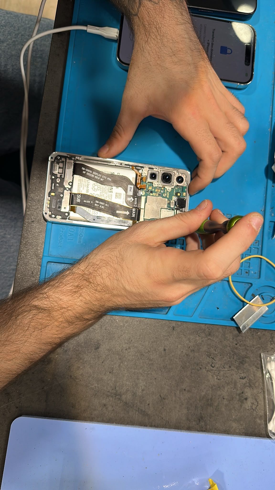 Des mains réparent un smartphone sur un tapis de travail bleu, avec des outils à proximité.