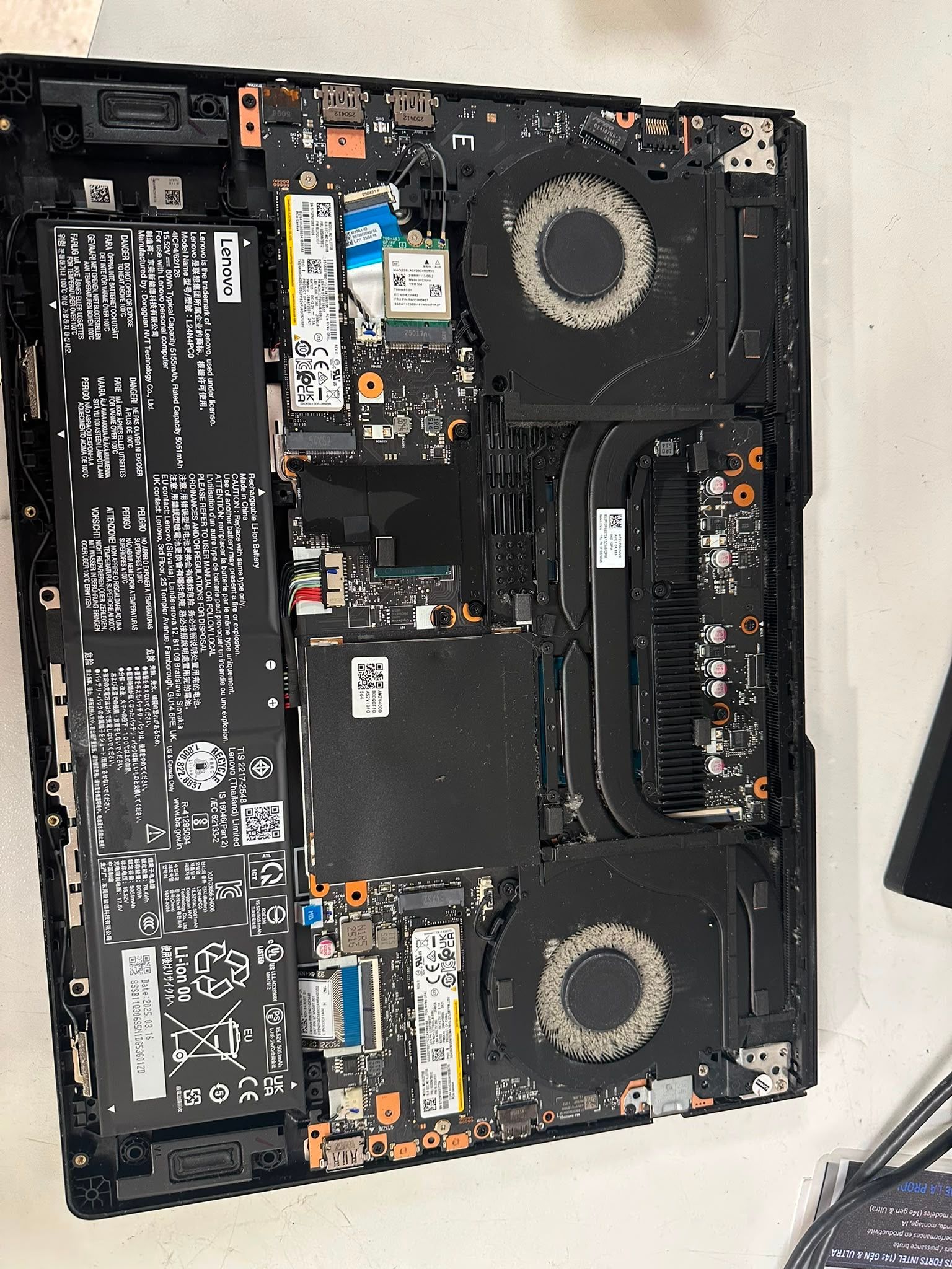 Intérieur d'un ordinateur portable ouvert, laissant apparaître la batterie, les ventilateurs, la carte mère et les composants sur une surface blanche.
