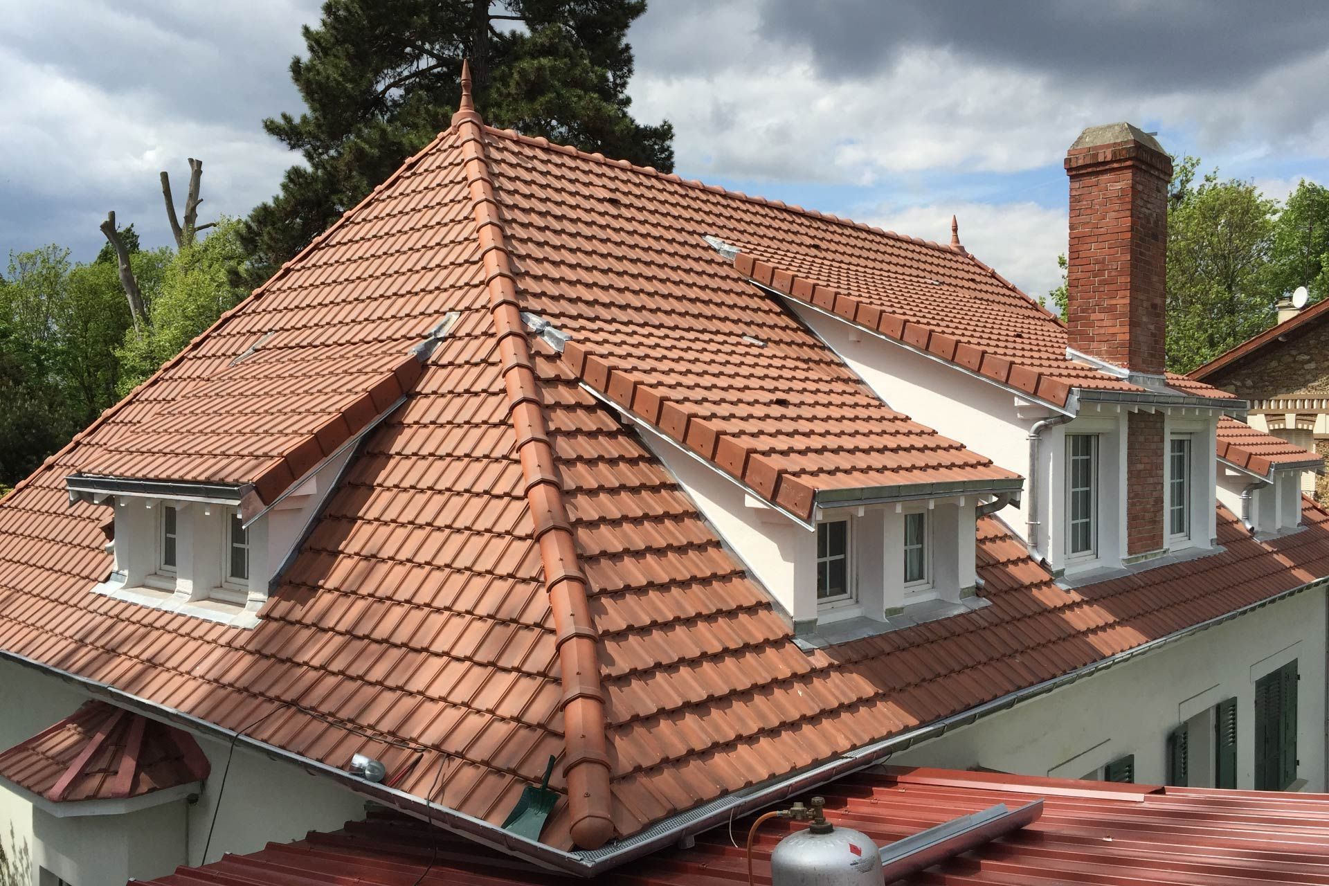 Maison au toit de tuiles rouges avec lucarnes et cheminée en briques sous un ciel partiellement nuageux