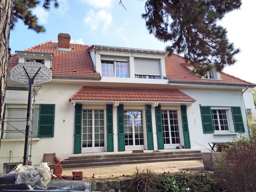 Maison blanche au toit de tuiles rouges, aux volets verts et au porche ombragé par de grands arbres