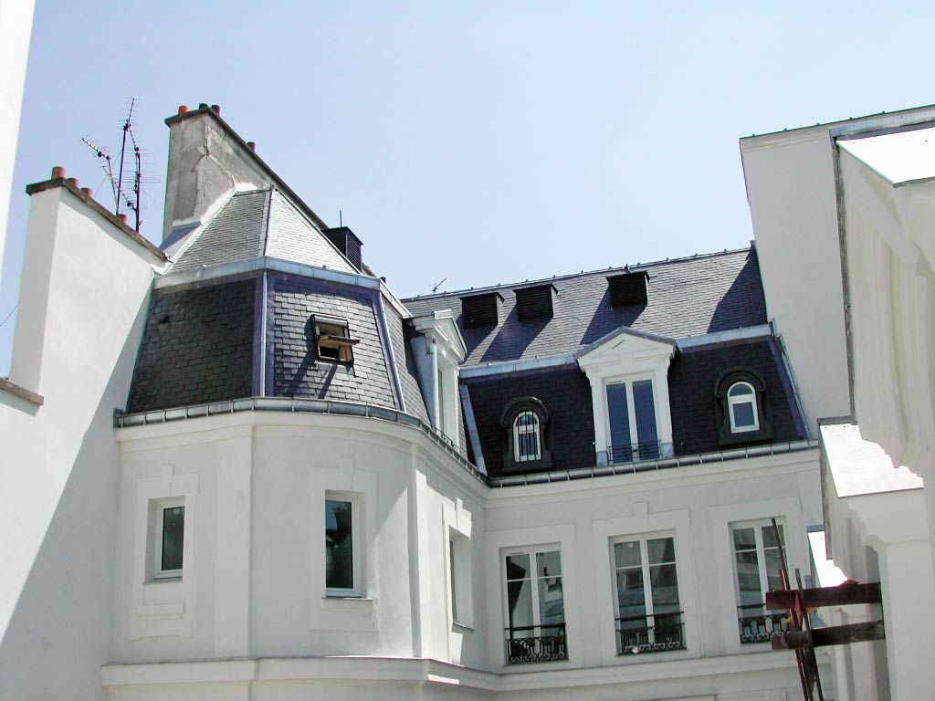 Bâtiment blanc avec un toit mansardé en ardoise sombre et des lucarnes sous un ciel lumineux