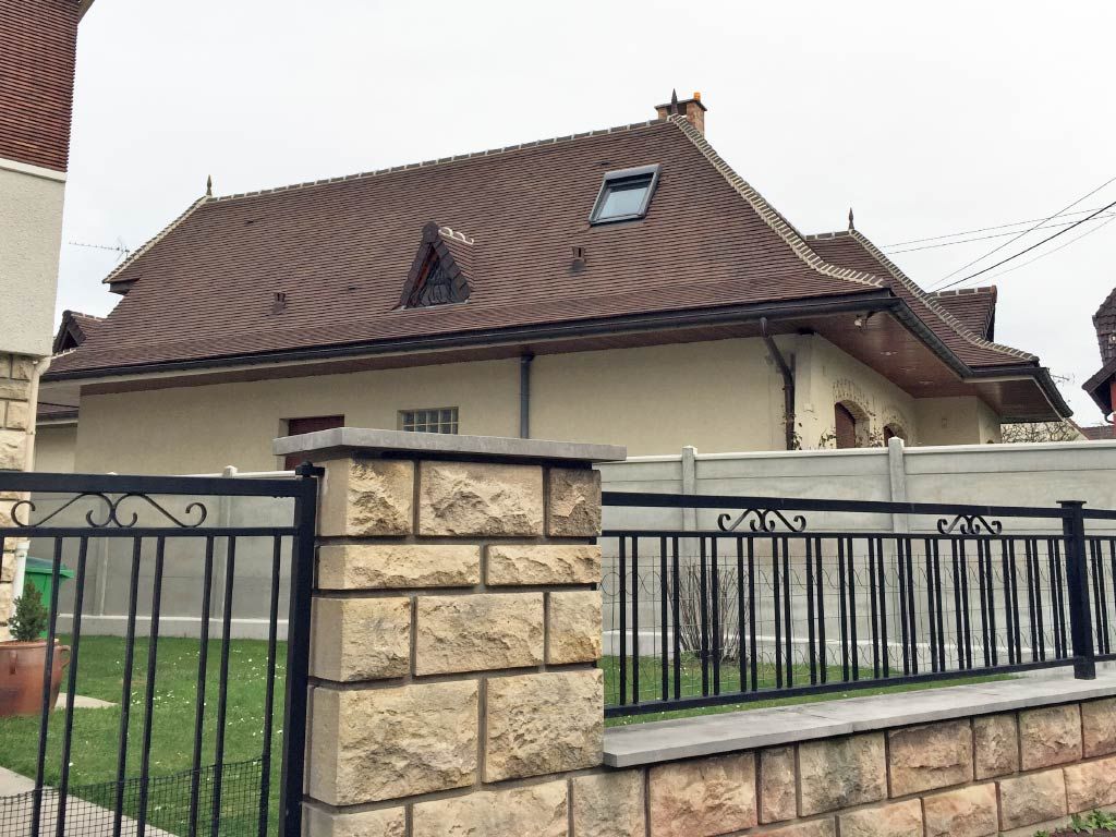 Maison individuelle au toit de tuiles brunes, derrière une clôture en pierre et en métal, par temps nuageux.