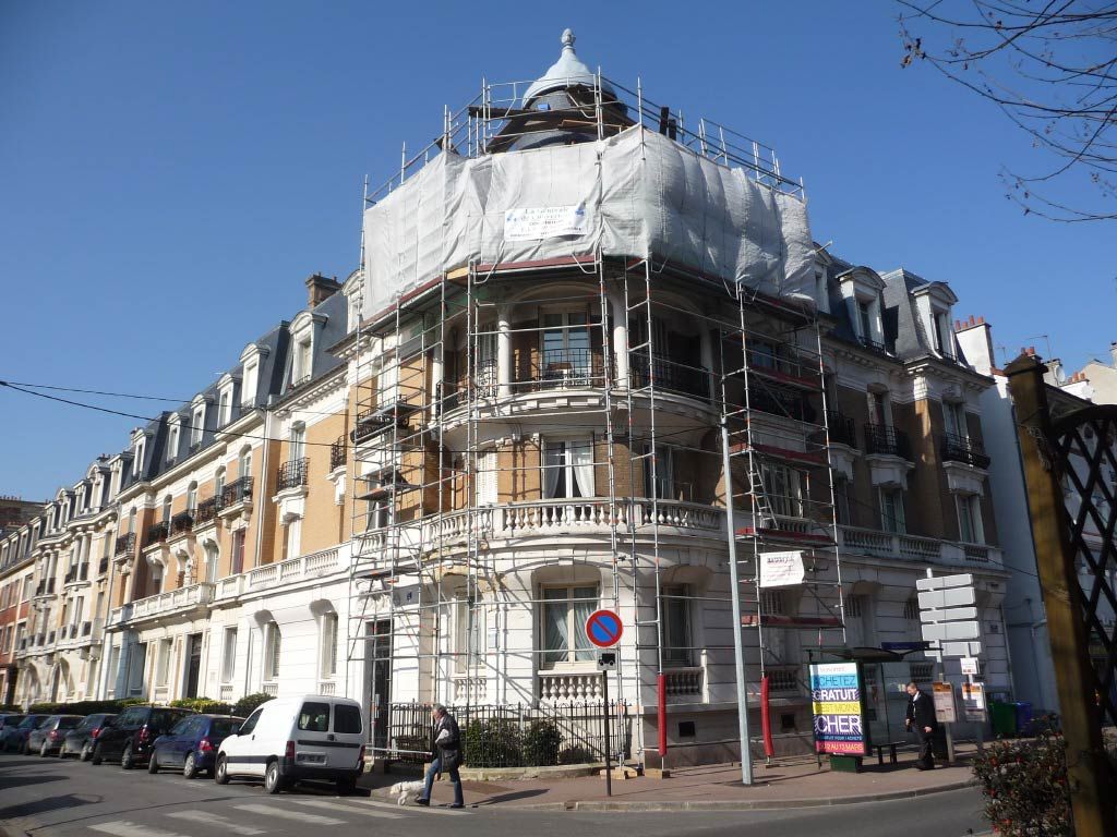 Immeuble d'angle en rénovation, recouvert d'échafaudages et de filets blancs, dans une rue ensoleillée.
