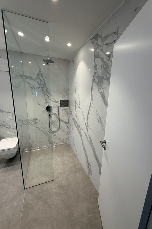 Modernes Badezimmer mit Dusche mit Fliesen in Marmoroptik und Glastür, grauem Boden, weißer Tür rechts.