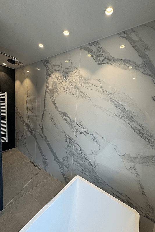 Modernes Badezimmer mit weißen, marmorähnlichen Wandfliesen, Deckenleuchten und einer weißen Badewanne.