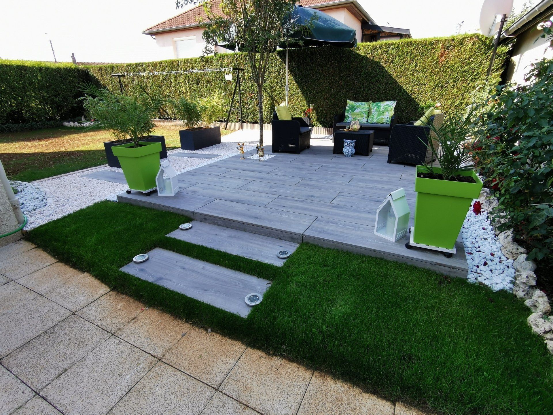 Une terrasse paysagée à l'arrière, des jardinières vertes, du gazon artificiel et des pierres décoratives blanches.