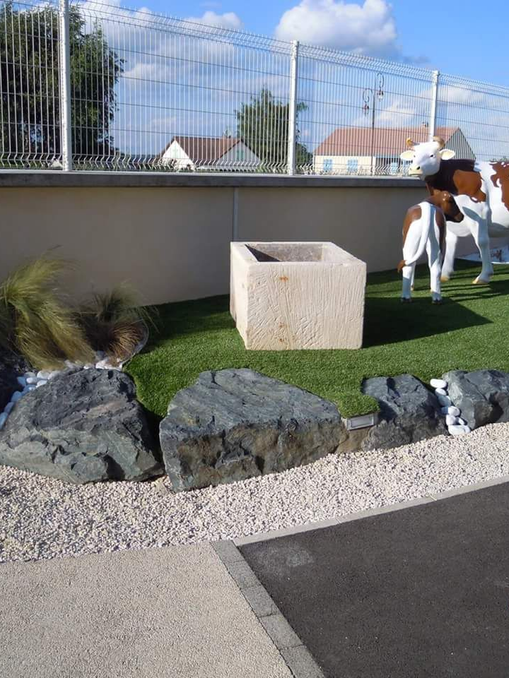 Un bac à fleurs carré en pierre est posé sur un carré de gazon artificiel devant une clôture, à côté d'une statue décorative de vache.