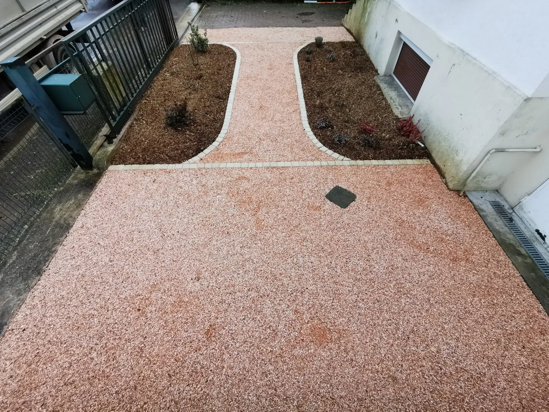 Un chemin pavé de granulats clairs mène à une porte, flanquée de deux parterres de jardin paillés à côté d'un bâtiment.