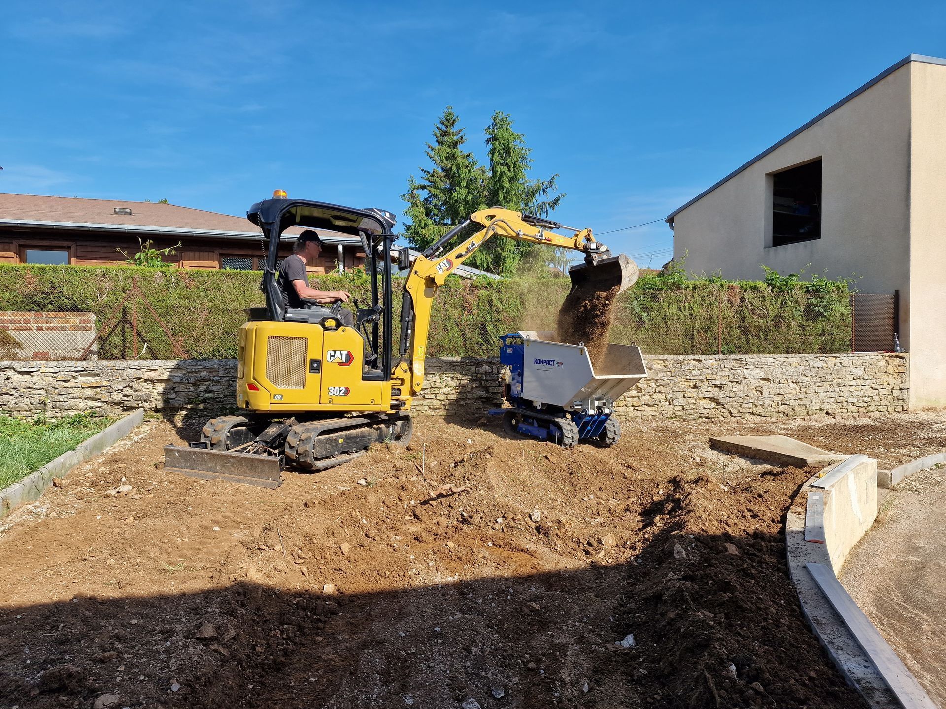 Une pelle mécanique Caterpillar jaune déverse de la terre dans un petit camion-benne chenillé sur un chantier de construction situé dans une cour résidentielle.