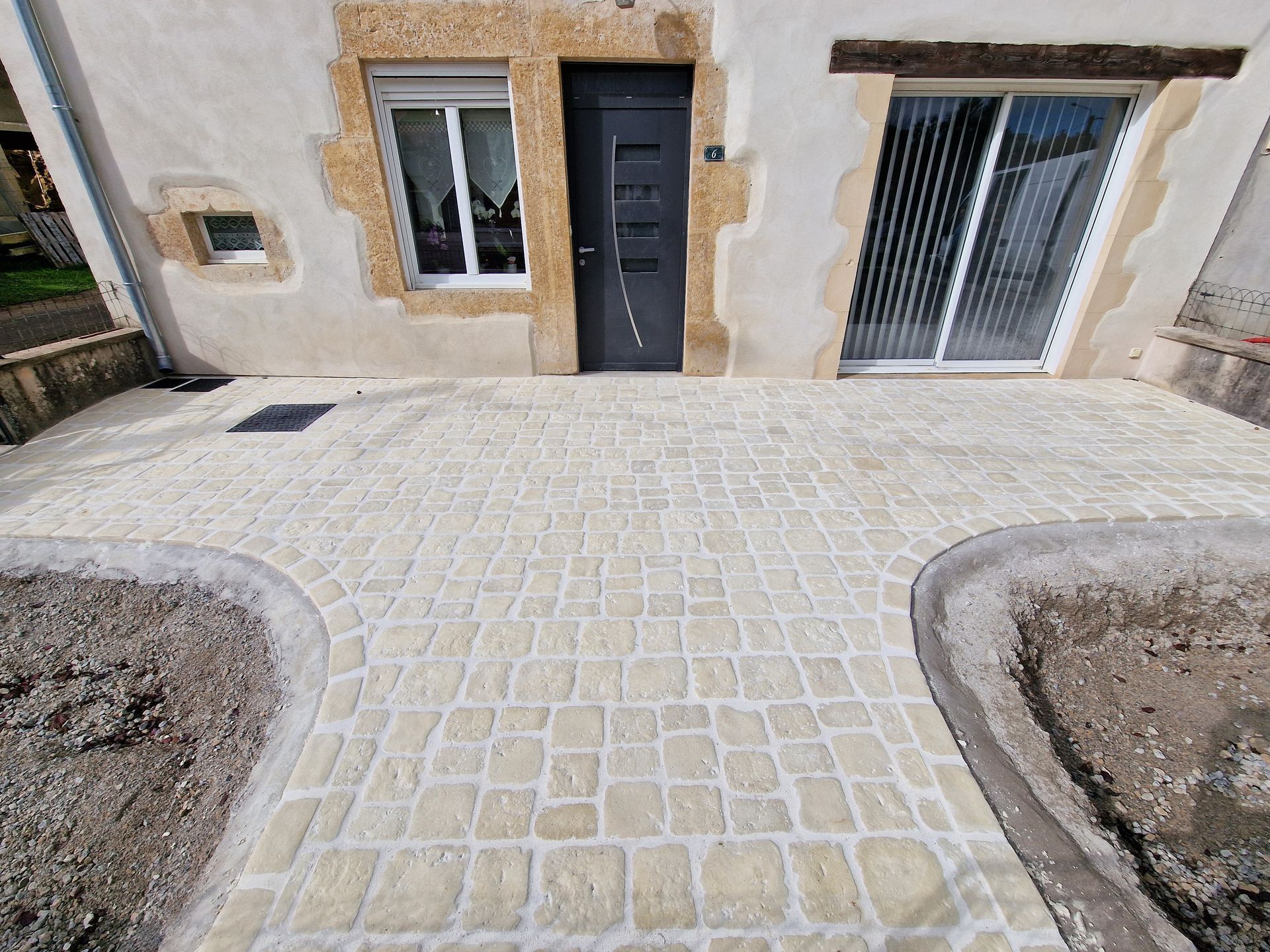 Une terrasse pavée de pierres claires, agrémentée de parterres de jardin en arc de cercle, mène à l'entrée d'une maison aux murs ornés de pierres.