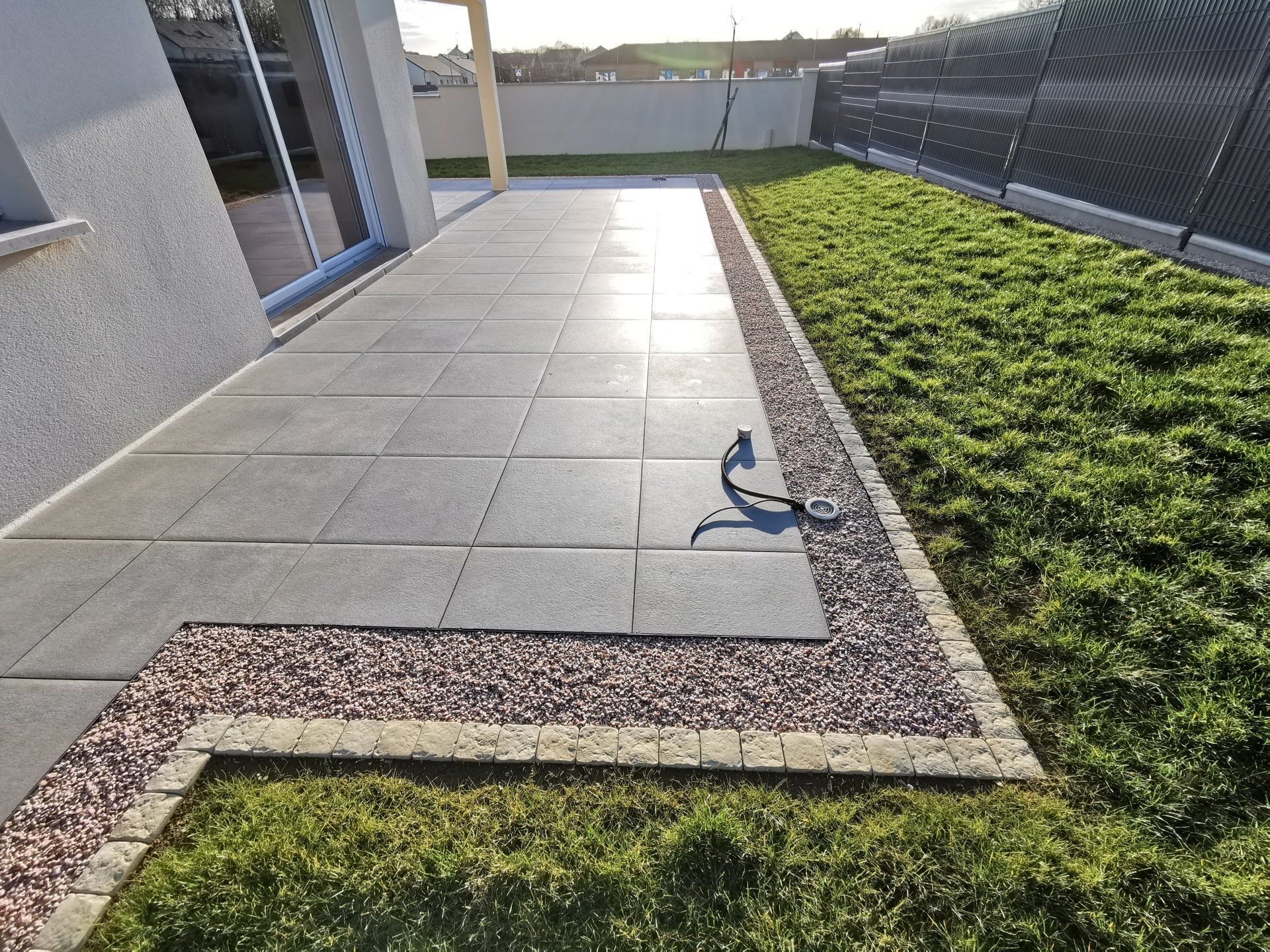 Une terrasse carrelée, bordée d'une bande de gravier et d'une bordure en pierre, jouxte une pelouse verte.