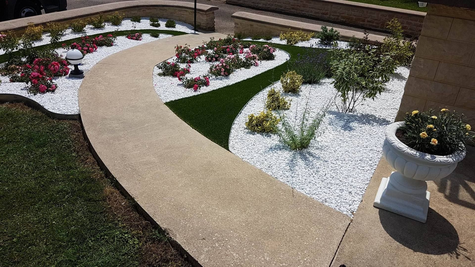 Une allée en béton incurvée traverse un jardin agrémenté de gravier blanc, de touffes de gazon vert, de petits arbustes et d'une jardinière blanche.