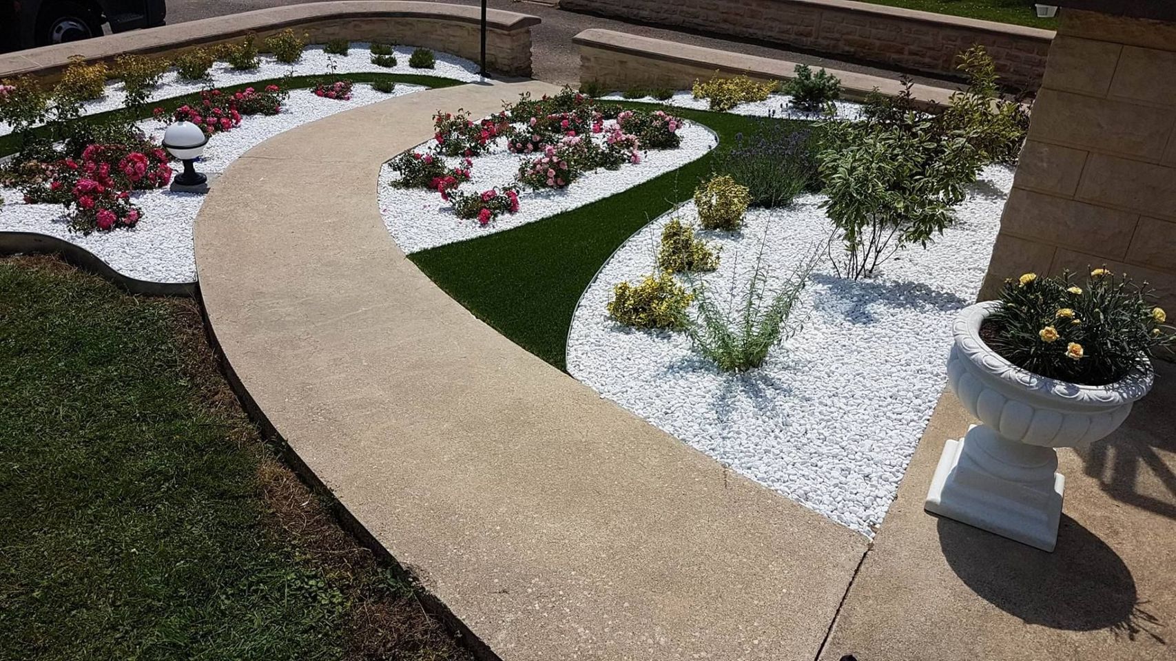 Un chemin de pierre sinueux serpente à travers un jardin paysager agrémenté de parterres de gravier blanc, de petits arbustes verts, de fleurs et d'une jardinière.