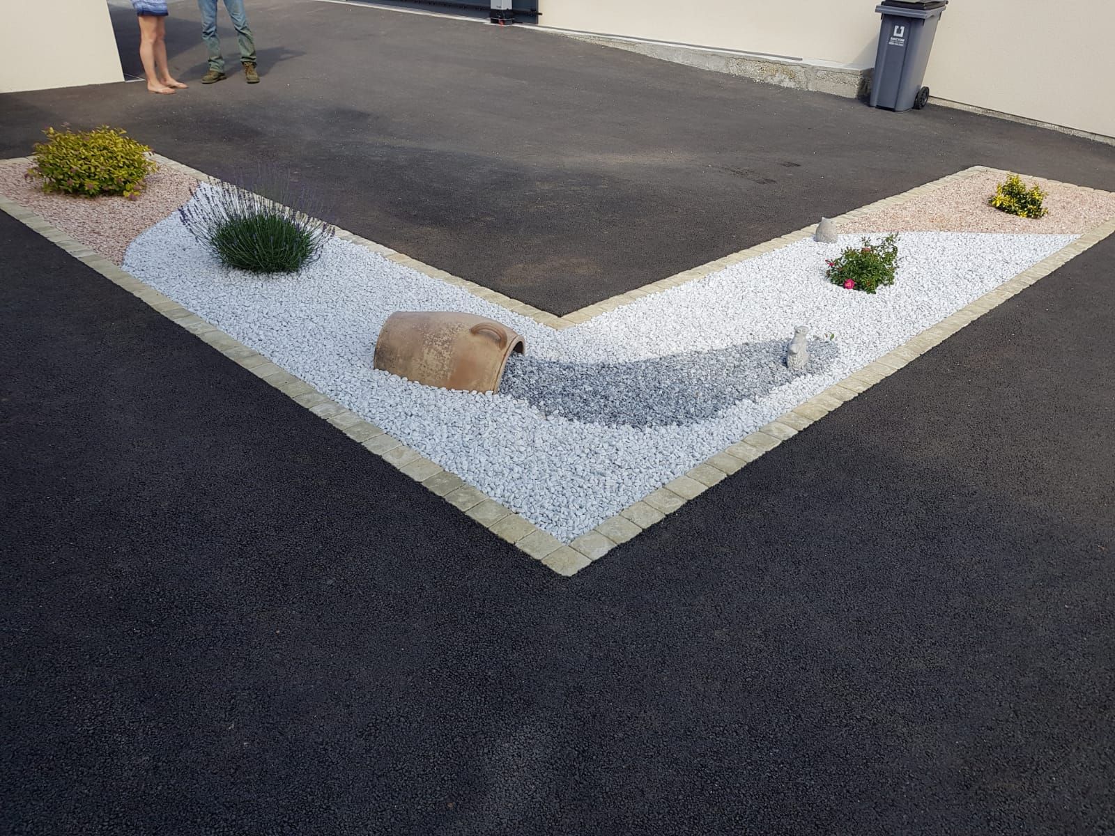 Un parterre de jardin en forme de L, recouvert de gravier blanc, agrémenté de roches décoratives et de petites plantes, bordant une allée pavée.