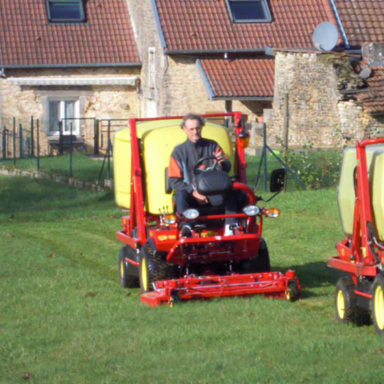 Une personne conduit une tondeuse à gazon rouge vif avec un réservoir jaune sur une pelouse herbeuse devant des maisons rustiques en pierre.