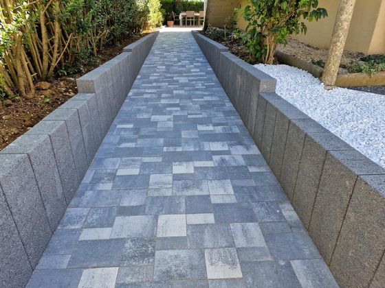Une allée pavée ornée d'un motif gris de différentes tailles, encadrée par de bas murets de pierre et du gravier blanc.
