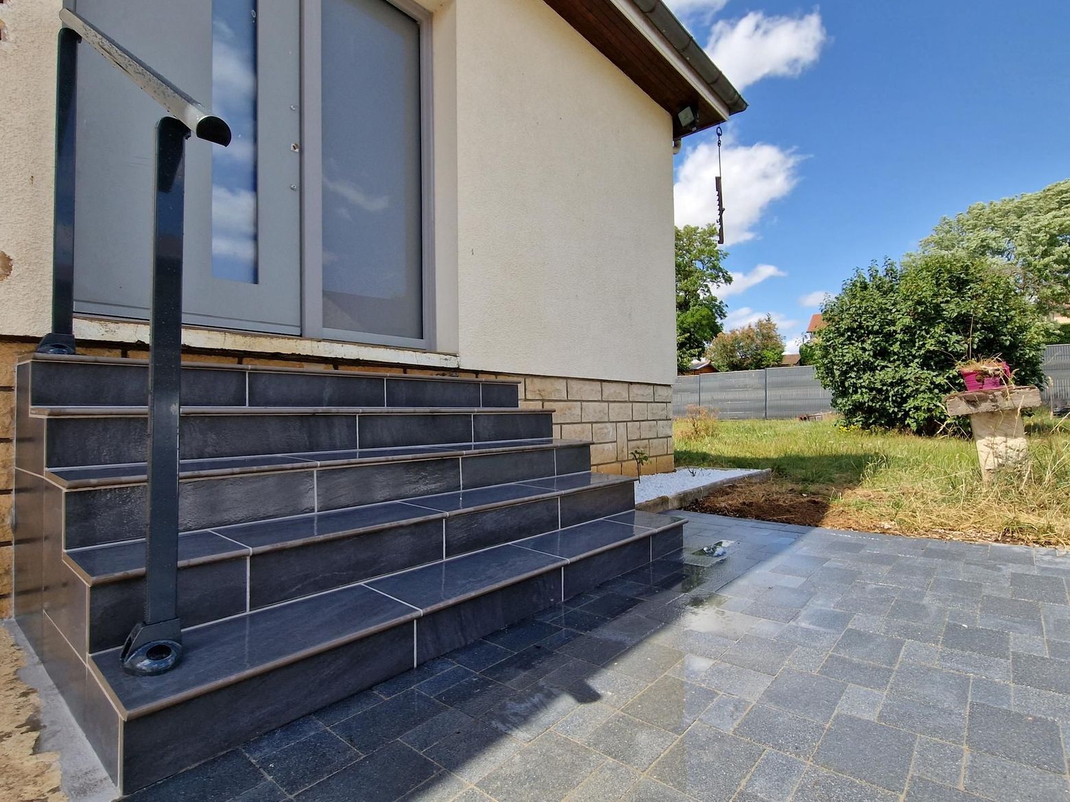 Des marches extérieures carrelées de couleur sombre, avec une rampe en métal noir, mènent à une porte-fenêtre.