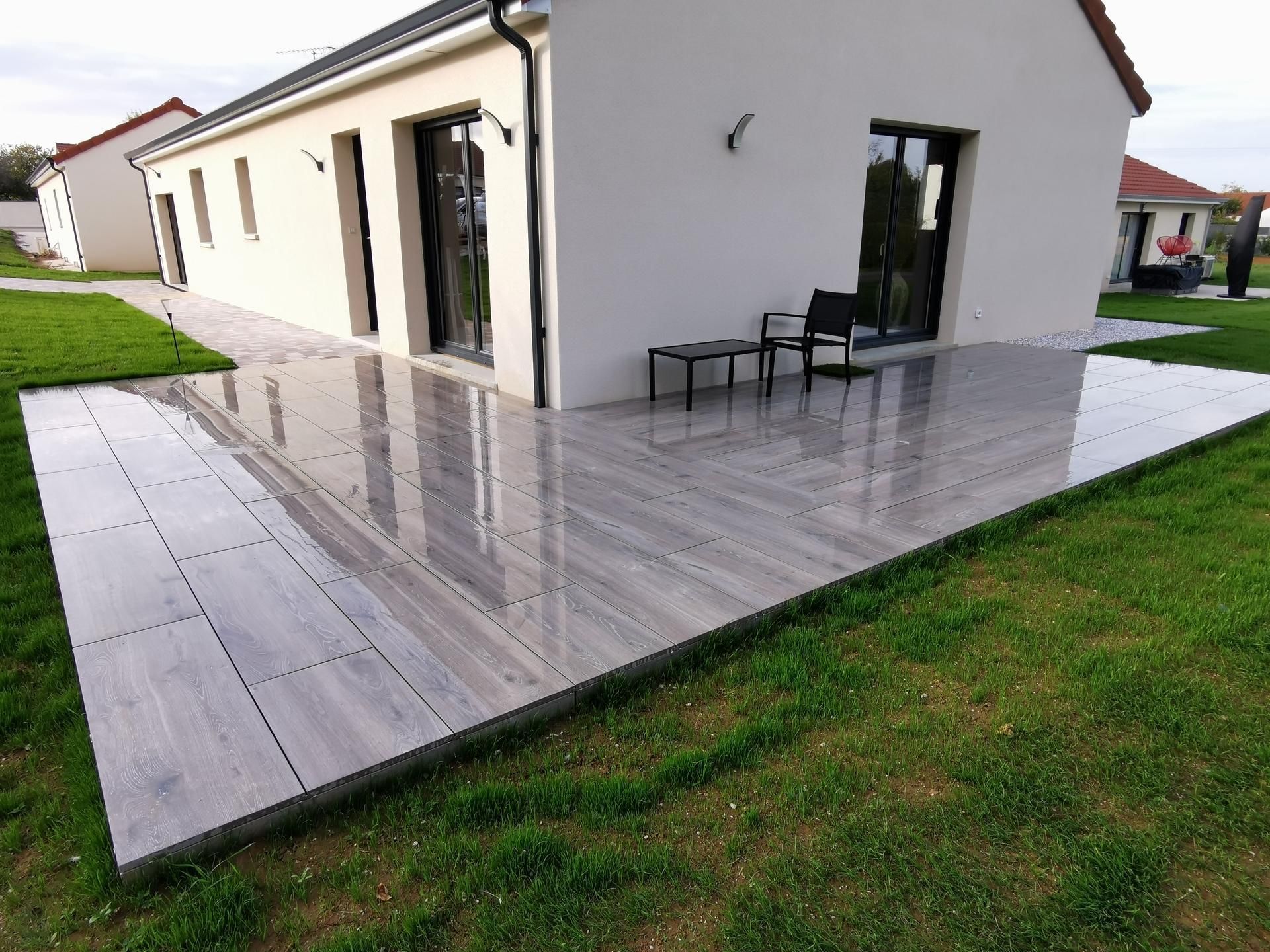 Une terrasse extérieure moderne, carrelée de gris, bordant une maison blanche, avec une petite table et une chaise placées contre le mur.