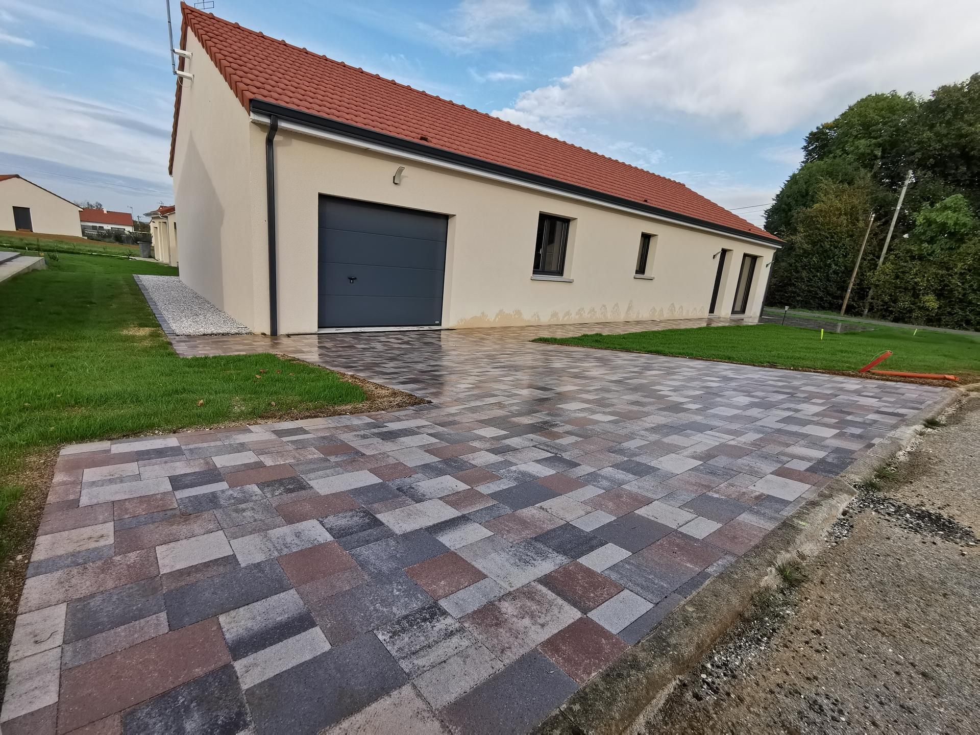 Une maison moderne de plain-pied, de couleur beige, avec un toit rouge et une allée pavée multicolore devant un garage.