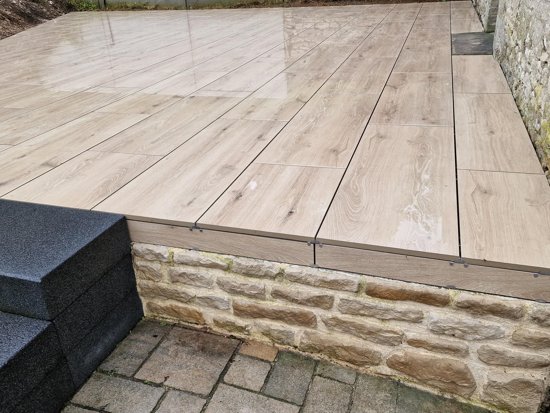 Une terrasse avec des carreaux imitation bois clair, un muret en pierre surélevé et des marches en pierre gris foncé au premier plan.