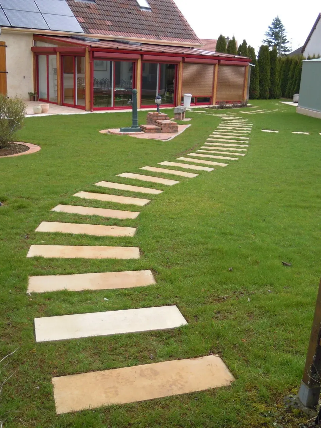 Un chemin sinueux de dalles de pierre rectangulaires, aménagé dans une pelouse verdoyante, mène à une maison dotée d'une véranda aux cadres rouges.
