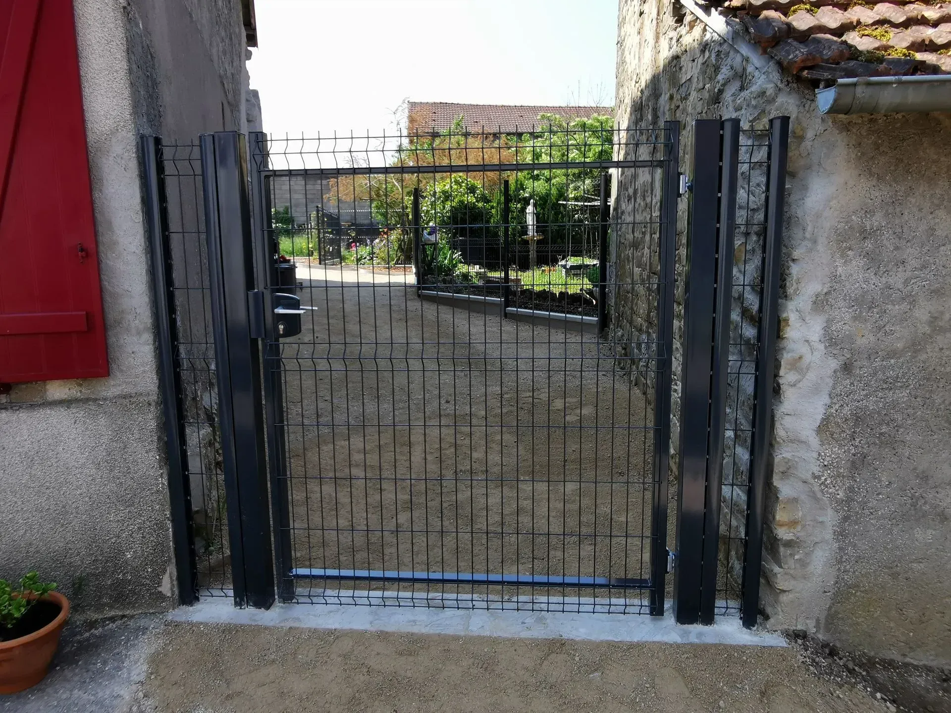 Un portillon piétonnier en métal noir se dresse entre deux murs d'immeuble en pierre donnant sur un chemin de jardin en gravier.