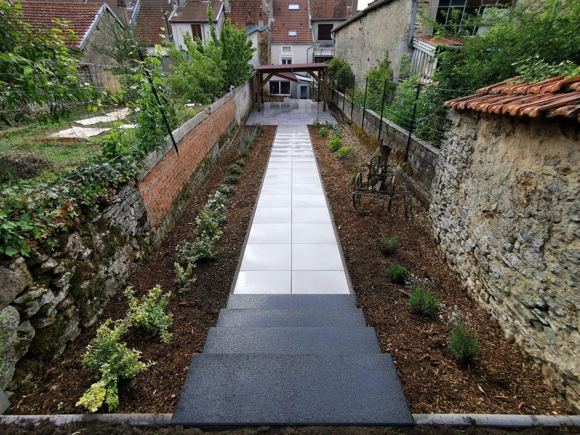 Un étroit chemin de pierre mène à un patio couvert, bordé de parterres de paillis avec de petites plantes entre deux vieux murs de pierre.