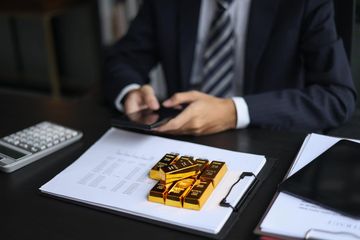 Empresario sentado en su escritorio con calculadora, teléfono y portapapeles sosteniendo lingotes de oro