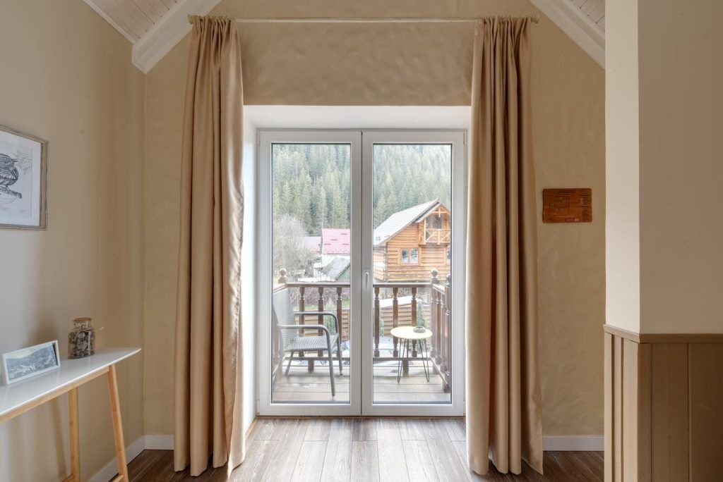 Intérieur d'une chambre avec des portes-fenêtres ouvertes donnant sur un balcon en bois, offrant une vue sur des chalets et une forêt en arrière-plan.