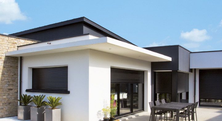Maison moderne avec des fenêtres et volets roulants gris foncés devant une terrasse